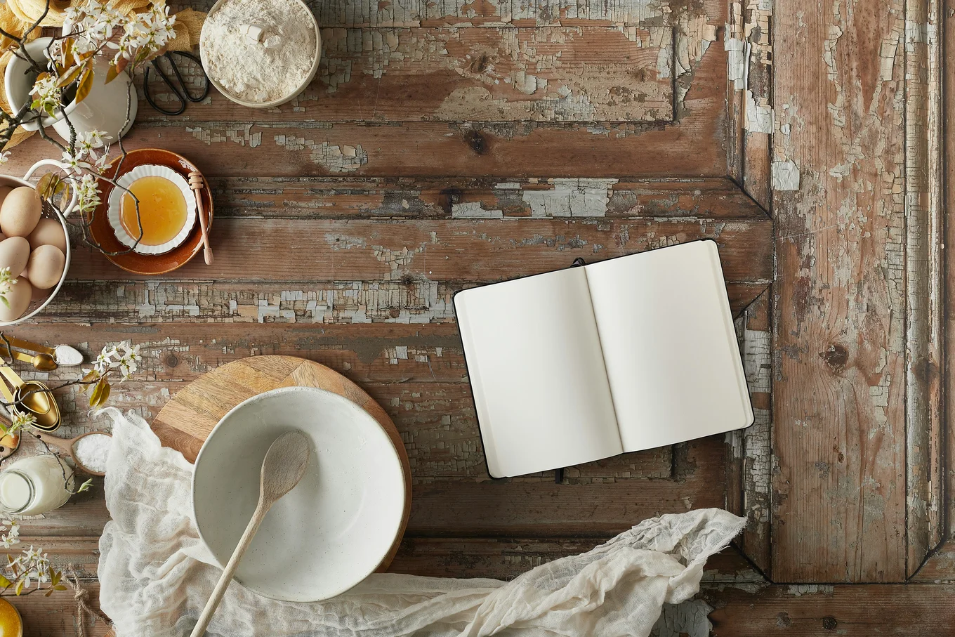 Top-down view of cooking utensils on a distressed wooden surface, including a bowl, wooden spoon, cutting board, bowl of flour, bowl of honey, milk, eggs, and a tea towel.